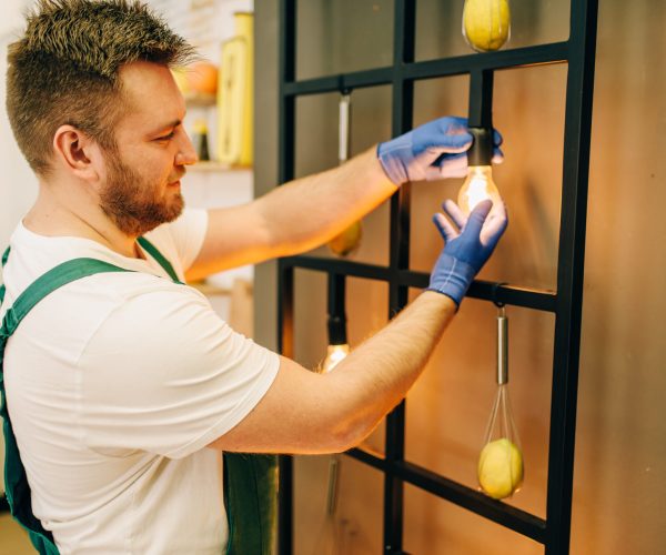 Electrician in uniform changes the light bulb, handyman. Professional worker makes repairs around the house, home repairing service