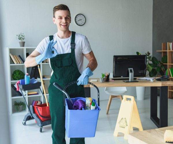 handsome smiling young cleaner holding bucket with cleaning supplies and showing ok sign
