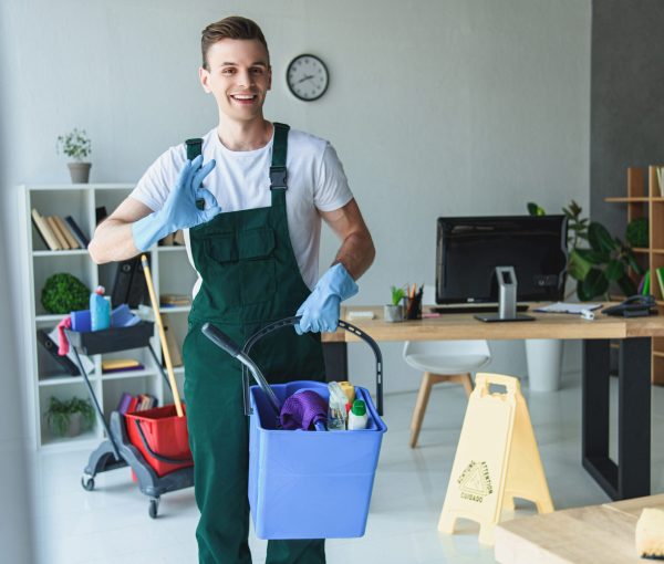 handsome smiling young cleaner holding bucket with cleaning supplies and showing ok sign