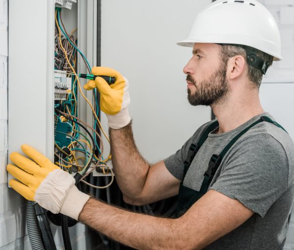 side view of handsome bearded electrician repairing electrical box and using screwdriver in corridor