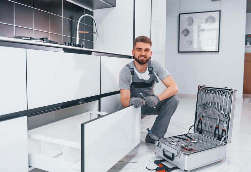 Young professional plumber in grey uniform using equipment on the kitchen and preparing for the work.