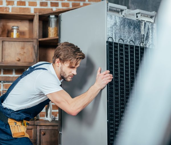 young repairman in protective workwear moving refrigerator in kitchen