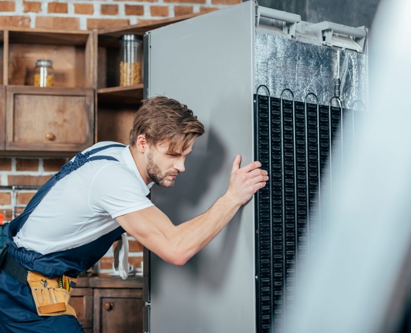 young repairman in protective workwear moving refrigerator in kitchen